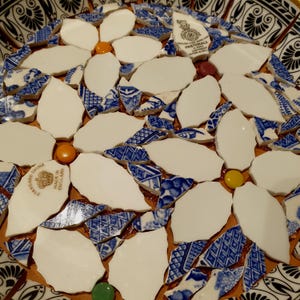 May include: A mosaic art piece crafted from broken ceramic shards featuring white petal shapes and blue and white patterned fragments. Small orange, yellow, green, and maroon circular beads are interspersed among the ceramic pieces.