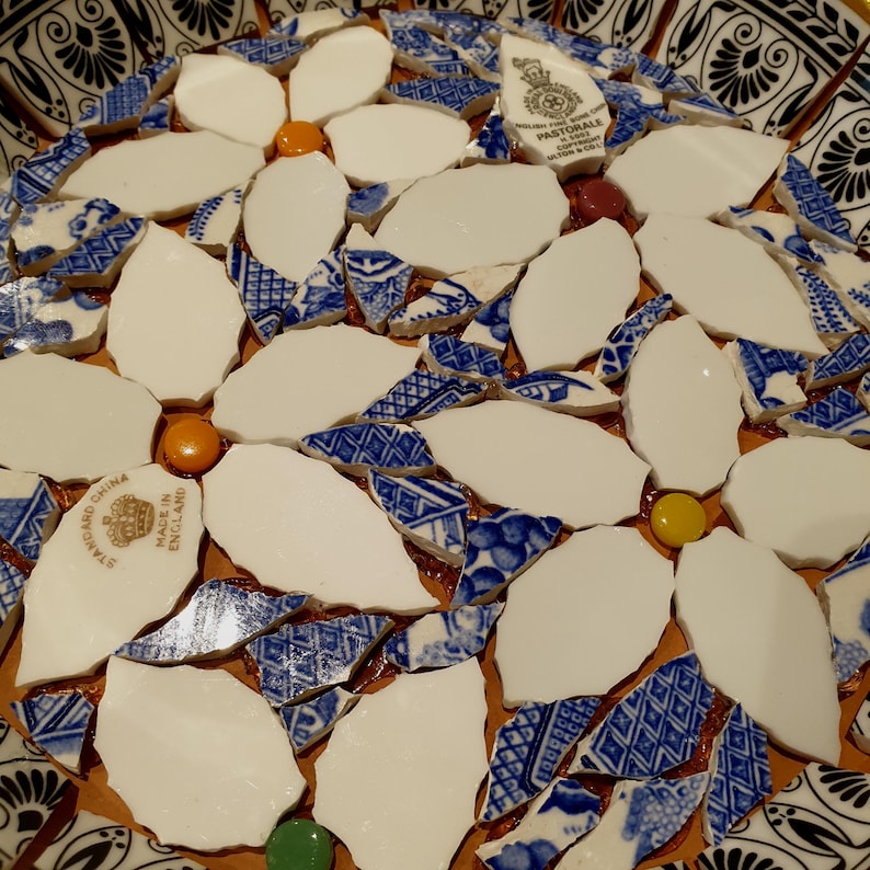 May include: A mosaic art piece crafted from broken ceramic shards featuring white petal shapes and blue and white patterned fragments. Small orange, yellow, green, and maroon circular beads are interspersed among the ceramic pieces.