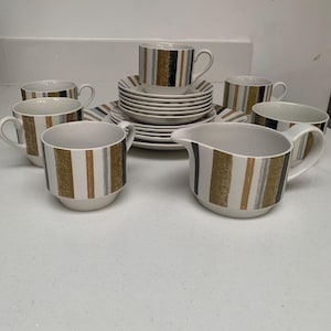 Op de afbeelding: Een wit keramisch theeservies met een goud, zwart en grijs gestreept ontwerp. De set bevat kopjes, schotels en een melkkan. De kopjes hebben oren en de schotels zijn rond. De melkkan heeft een tuit en een oor.