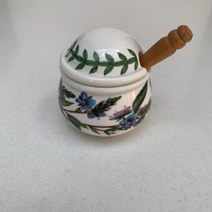 May include: A white ceramic sugar bowl with a lid and a wooden spoon. The bowl is decorated with green leaves and blue and purple flowers. The spoon is made of light brown wood.