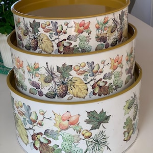 Pode incluir: Um conjunto de três latas de armazenamento redondas e empilháveis com fundo branco e uma estampa botânica de bolotas, pinhas, roseiras e várias folhas de outono. As bordas das latas são de uma cor amarelo-mostarda sólida.