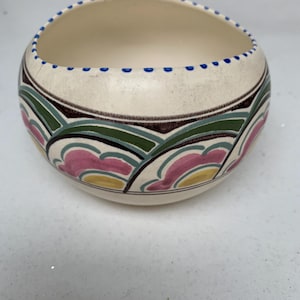 May include: A cream-colored ceramic bowl with a rounded shape. The bowl features a decorative pattern of pink, green, and yellow shapes, outlined in brown and blue. The rim is accented with small blue dots.