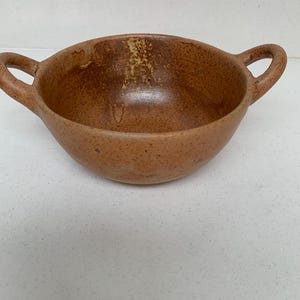 May include: A small, round, brown ceramic bowl with two loop handles. The bowl has a speckled texture and a slightly irregular shape. The interior has a lighter brown colour with some darker spots.