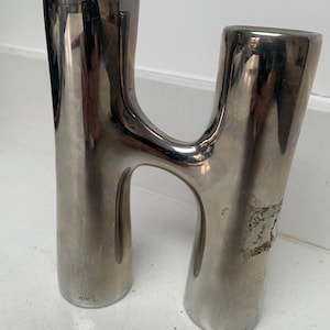May include: A silver-toned metal sculpture in the shape of the letter "H". The sculpture has a smooth, reflective surface with some wear and tear. The base is slightly wider than the top, and the piece is free-standing.