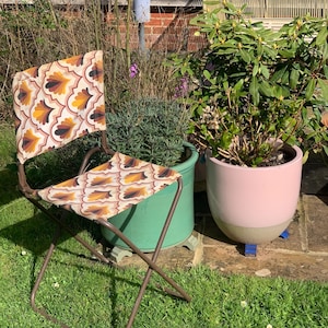 Peut inclure: Chaise pliante d'extérieur avec une structure en métal marron et une assise et un dossier en tissu à motifs. Le tissu présente un motif répétitif dans les tons marron, jaune et crème. La chaise est posée sur une pelouse.