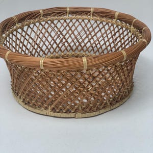May include: A round, open-top basket constructed from brown pine needles and light-colored raffia or grass. The sides feature a diagonal lattice pattern, while the rim and base are reinforced with thicker, coiled bundles of pine needles.