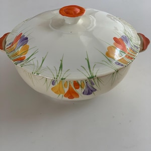 May include: A white ceramic serving dish with a lid, featuring a floral design in orange, yellow, and purple. The dish has orange handles and a matching orange knob on the lid. The floral pattern is complemented by green grass-like stems.