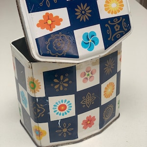 May include: A hexagonal metal storage tin featuring a checkerboard pattern of dark blue squares and white squares. The white squares are decorated with various colourful floral and geometric motifs in shades of orange, yellow, pink, and blue.