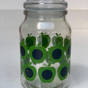 May include: A clear glass storage jar with a wide mouth and a matching glass lid. The exterior of the jar is decorated with a repeating pattern of bright green apple shapes, each featuring a solid dark navy blue circle in the centre.