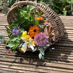 May include: A rustic wicker basket brimming with a colourful array of fresh flowers and foliage. The arrangement features orange, yellow, purple, and white flowers, alongside verdant green leaves. The basket rests on a wooden slatted surface.