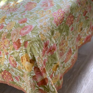 Op de afbeelding: Een stoffen sprei met een dicht bloemenpatroon van roze, gele en oranje bloesems op een witte achtergrond met groen blad. De randen zijn afgewerkt met een effen perzikkleurige bies.