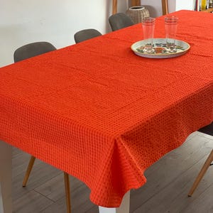 May include: A bright orange rectangular tablecloth with a textured waffle weave pattern draped over a dining table. On the surface sits a round tray containing two clear glass tumblers with horizontal gold rim details near the top edges.