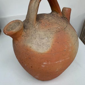 May include: A rustic, terracotta-colored jug with a large handle and two spouts. The jug has a rounded body and shows signs of age, with a weathered appearance. The handle is arched, and the spouts are cylindrical. The jug is likely handmade.