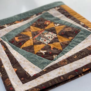May include: A quilted fabric piece featuring a central star pattern in mustard yellow and dark brown. The quilt includes borders of sage green with leaf patterns, cream fabric with small floral motifs, and dark brown fabric with cat illustrations.