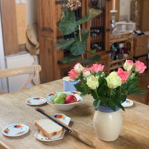 Puede incluir: Una mesa de madera está decorada con un ramo de rosas rosas y amarillas en un jarrón crema con borde azul. También hay pequeños platos con diseños coloridos, un cuchillo para pan y un cuenco de fruta.