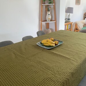 May include: A rectangular dining table covered with a textured olive green waffle-weave tablecloth. Centered on the table is a square ceramic plate featuring a green and white pattern, holding a cluster of yellow bananas and two red apples.