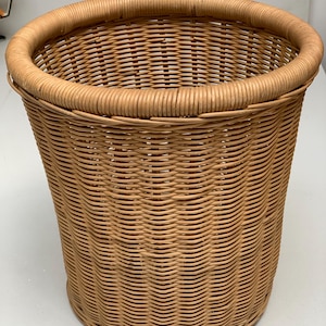 Peut inclure: Une corbeille à papier cylindrique fabriquée en osier tressé de couleur brun clair. Le panier présente un bord supérieur épais et arrondi ainsi qu'un motif de tressage serré et régulier sur tout le corps, formant un récipient ouvert robuste.