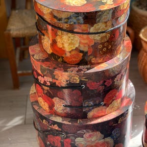 Puede incluir: Una pila de tres cajas redondas con tapa que presentan un fondo oscuro con un vibrante estampado floral de flores rojas, naranjas, amarillas y blancas. Cada caja tiene un asa de cordón negro fijada en el lateral.