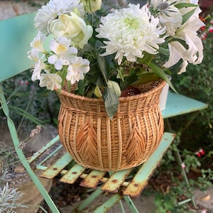 Può includere: Un cesto rotondo in vimini intrecciato color marrone chiaro con bordo svasato e un motivo decorativo a canne verticali. Il cesto è riempito con una rigogliosa composizione di crisantemi bianchi, rose bianche, gigli bianchi e fiori di cardo blu.