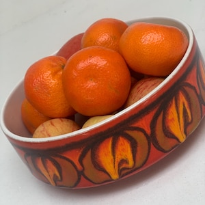 Könnte beinhalten: Eine runde Keramikschale mit weißem Innenraum und einer leuchtend orangefarbenen Außenseite, die ein sich wiederholendes, abstraktes, flammenartiges Muster in Dunkelbraun und Gelb aufweist. Die Schale ist mit Mandarinen und zwei Äpfeln gefüllt.