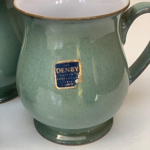 May include: A sage green ceramic mug with a white interior and a curved handle. The mug features a blue square sticker on the front with the text "THE DENBY POTTERY COMPANY LTD" in gold lettering. The exterior has a speckled finish and a base.