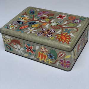 May include: A rectangular metal storage tin with a light olive green base colour. The lid and sides are decorated with a vibrant, multi-coloured folk-art style floral pattern featuring red, orange, yellow, blue, pink, and white painted motifs.
