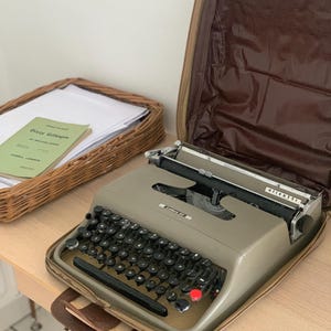 May include: A beige Olivetti Lettera 22 portable typewriter with a black keyboard and a red key, housed in its open brown carrying case. Beside it sits a wicker basket containing white papers and a book titled Dizzy Gillespie by Michael James.