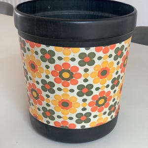 May include: A cylindrical waste bin featuring a black plastic rim and base. The central section is decorated with a repeating pattern of stylised flowers in shades of orange, yellow, and olive green against a white background, creating a vibrant look.