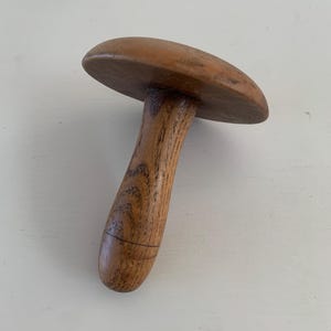 Puede incluir: Objeto de madera en forma de hongo con una parte superior redondeada y un mango curvo. La veta de la madera es visible y el color es marrón cálido. El objeto parece ser una herramienta o un elemento decorativo.