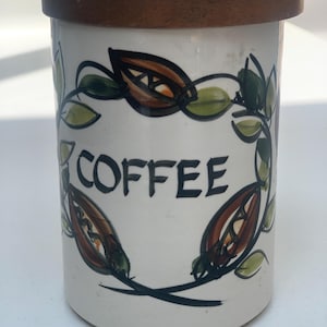 Puede incluir: Un bote de café cilíndrico de cerámica blanca con un diseño pintado a mano de hojas verdes y granos de café marrones que rodean la palabra COFFEE en letras negras. El recipiente está rematado con una tapa plana de madera marrón oscuro.