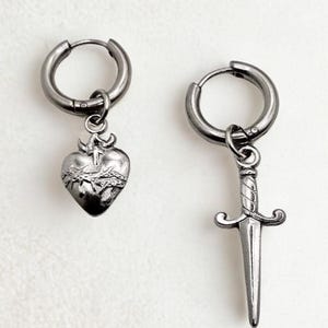 Puede incluir: Dos pendientes de aro plateados. Un pendiente presenta un colgante de corazón con una corona de espinas y una cruz. El otro pendiente tiene un colgante de daga. Los pendientes se muestran sobre un fondo blanco.