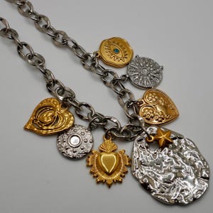 Puede incluir: Un collar de cadena de plata con múltiples dijes, incluyendo corazones de oro, un corazón de oro con un sol radiante, un sol radiante de plata, un ojo de plata con una piedra azul, un disco de plata con una estrella y un disco de plata con un diseño grabado.