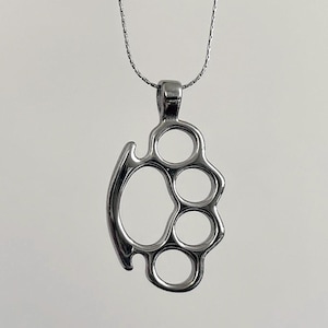 May include: A silver pendant necklace with a knuckle duster design. The pendant has four finger holes and a curved handle. The necklace chain is silver and hangs against a light background.