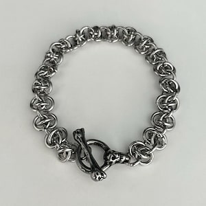 May include: A silver-toned chainmail bracelet with a toggle clasp. The clasp features a bone-shaped bar and a skull detail. The bracelet is made of interlocking metal rings.