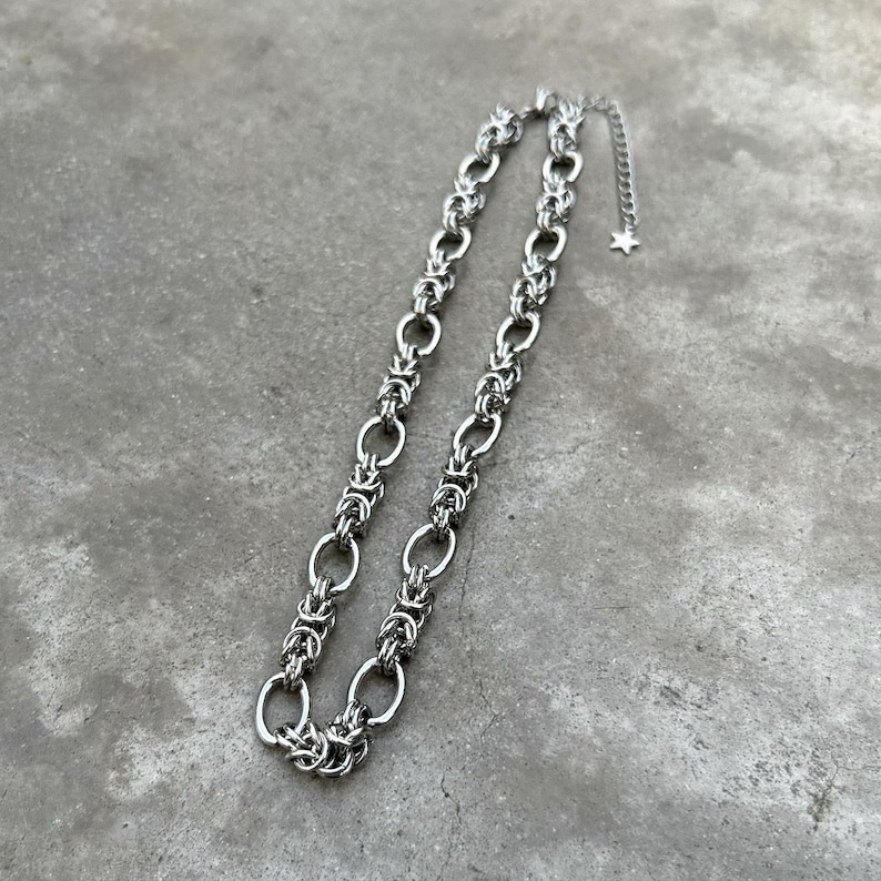 May include: A silver chain necklace with a delicate, intricate design. The chain is made of small, linked rings that create a woven pattern.