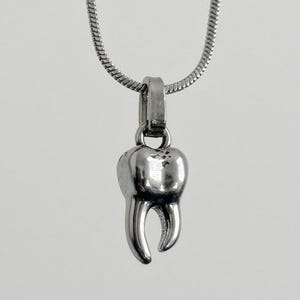 May include: A silver pendant necklace with a detailed tooth charm. The tooth charm has a realistic shape with roots and a crown. The necklace chain is a thin, textured silver. The background is a solid, neutral colour.