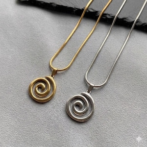 Puede incluir: Dos collares con colgantes en espiral. Uno es dorado, el otro plateado. Ambos collares tienen una cadena delicada y un colgante circular con un diseño en espiral. Los colgantes están centrados en una superficie gris.