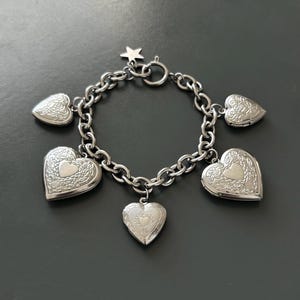 May include: Silver-toned charm bracelet featuring heart-shaped lockets and a star charm. The bracelet has a chain link design with six heart-shaped charms, each with intricate detailing. The bracelet is made of a shiny, metallic material.