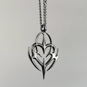 Puede incluir: Collar con colgante plateado con un diseño intrincado y simétrico. El colgante presenta una forma central en forma de corazón con elementos estilizados en forma de llama. La cadena es de estilo fino y enlazado.