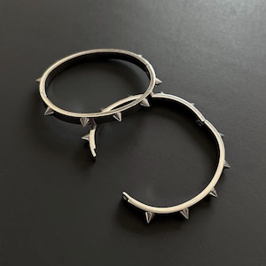 May include: Two silver-colored, spiked bracelets on a black surface. The bracelets are circular, with evenly spaced, pointed spikes around the outer edge. One bracelet is open, while the other is closed.