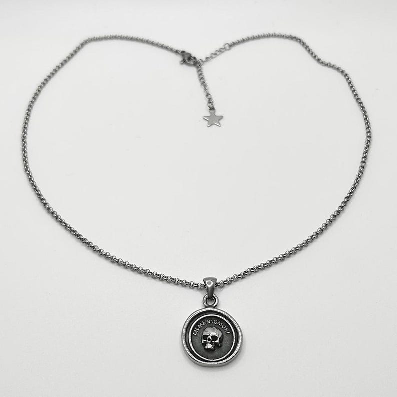 Op de afbeelding: Een zilverkleurige metalen ketting met een ronde hanger voorzien van een reli&euml;f van een schedel en de tekst MEMENTO MORI in reli&euml;f langs de rand. De ketting heeft een klassieke schakelstijl met een verstelbaar uiteinde met een sterretje.