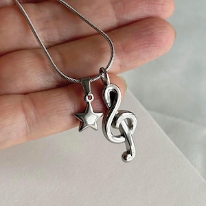 Peut inclure: Un collier en chaîne argentée avec deux pendentifs : une étoile et une clé de sol. Les pendentifs sont tous deux en métal et ont une finition polie.