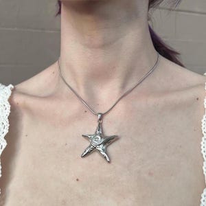 Puede incluir: Un collar de tono plateado con un colgante de estrella de mar de cinco puntas. El colgante tiene un acabado metálico liso con un diseño de espiral en relieve en el centro. La estrella de mar cuelga de una cadena de serpiente fina y flexible.
