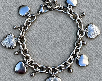 Puede incluir: Una pulsera de dijes plateada con un diseño de cadena de eslabones gruesos. La pulsera está adornada con dijes de corazón lisos y corazones con un intrincado patrón floral en relieve, intercalados con pequeñas esferas plateadas.