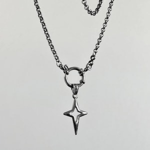 A silver-toned necklace featuring a four-pointed star pendant. The pendant hangs from a circular clasp, connected to a chain necklace. The chain has a classic link design. The jewelry is displayed against a neutral background.