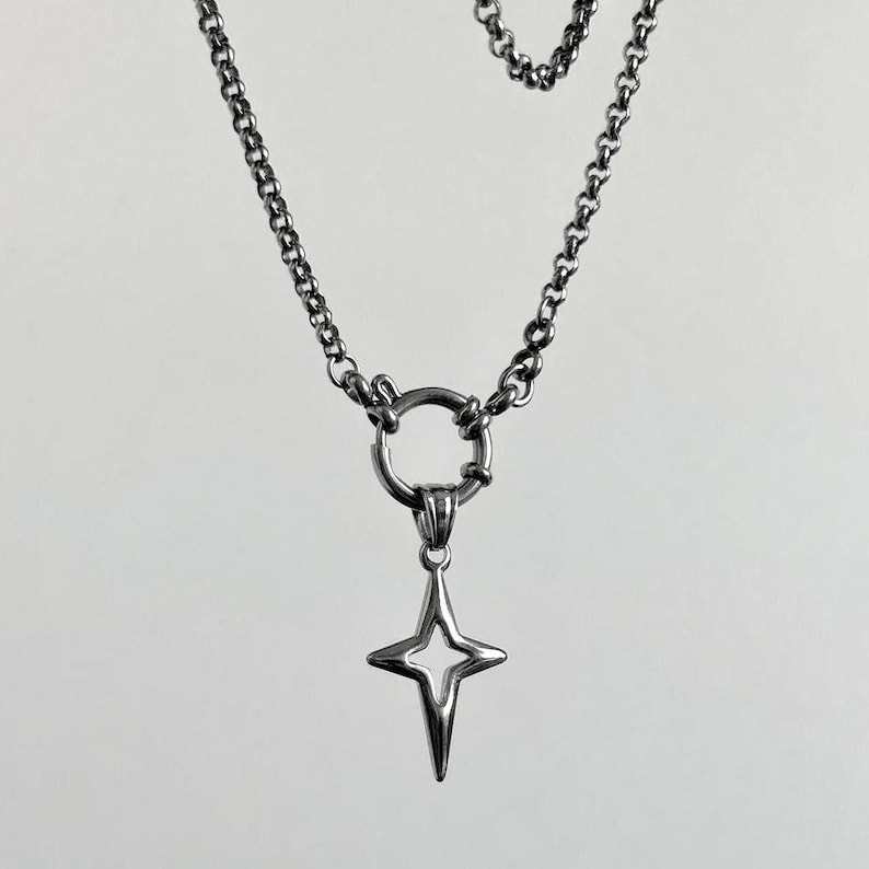 A silver-toned necklace featuring a four-pointed star pendant. The pendant hangs from a circular clasp, connected to a chain necklace. The chain has a classic link design. The jewelry is displayed against a neutral background.