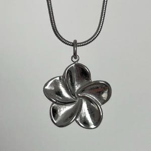 Könnte beinhalten: Halskette mit einem silberfarbenen Anhänger in Form einer fünfblättrigen Blume. Der Anhänger hängt an einer dünnen, strukturierten Kette. Die Blume hat eine polierte, reflektierende Oberfläche, vor neutralem Hintergrund.