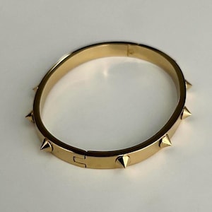 Puede incluir: Un brazalete dorado con una serie de púas puntiagudas espaciadas uniformemente. El brazalete tiene un cierre de bisagra y una superficie lisa y pulida. Las púas doradas añaden un elemento de diseño audaz y vanguardista. Circunferencia de unos 18 cm.