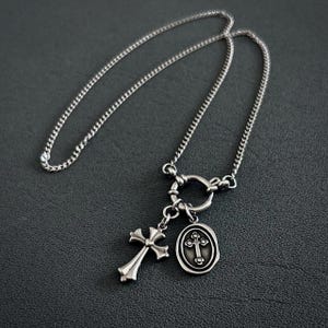Pode incluir: Um colar de corrente de prata com um pingente de cruz de prata e um pingente oval de prata com um design de cruz. O colar tem um fecho de trava.
