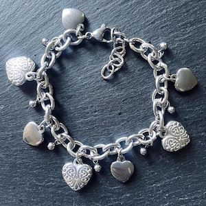 Puede incluir: Pulsera de abalorios plateada con un diseño de cadena gruesa. La pulsera está adornada con múltiples colgantes en forma de corazón, algunos con patrones intrincados y otros con un acabado liso. Una joya clásica.